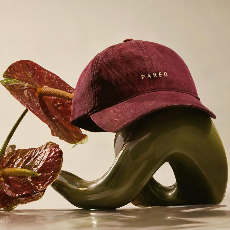 burgundy baseball cap fashion product photo: A burgundy embroidered cap rests on a glossy sculptural form beside a deep red flower, lit warmly against a pale s…
