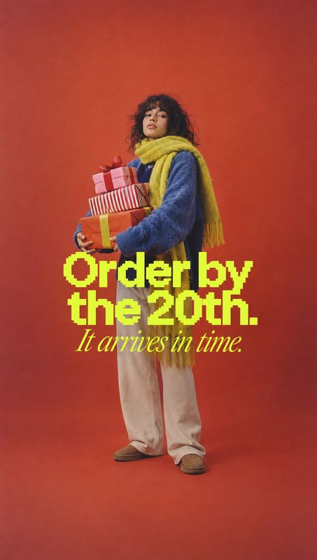 christmas product photo: A model in a blue sweater, yellow scarf and light trousers holds wrapped gifts against a vivid red studio backdrop, with bold yellow d…