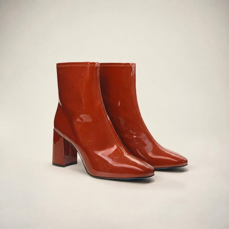 glossy ankle boots packshot product photo: A pair of glossy reddish-brown ankle boots with block heels is arranged against a softly lit off-white studio backdr…