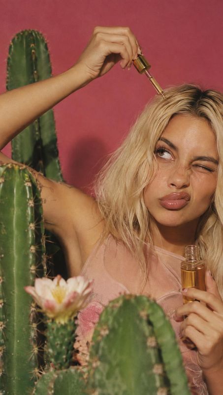 hair oil model try-on product photo: A blonde woman applies golden hair oil with a dropper among cacti against a warm pink studio backdrop, with playful expres…