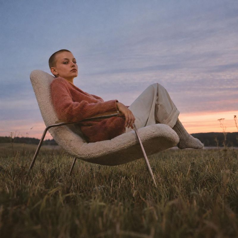 upholstered lounge chair lifestyle scene product photo: A close-cropped-haired woman in a rust-colored sweater, pale trousers, and socks reclines in a plush lo…