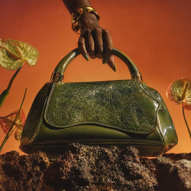 handbag product in hand product photo: A glossy dark green handbag is held by a hand wearing a gold cuff, staged among rugged rocks and translucent botanical f…