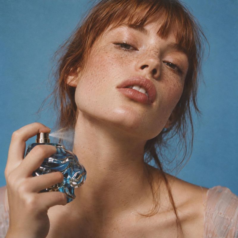 perfume beauty close-up product photo: A freckled woman holds a sculptural glass perfume bottle near her face against a blue studio backdrop, with soft light a…