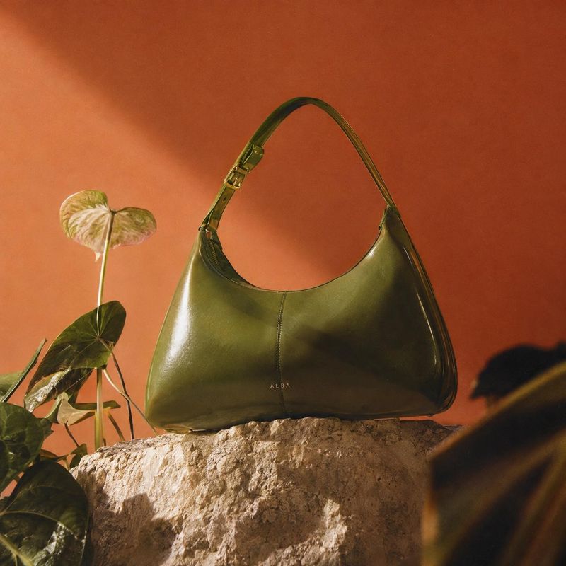 green leather handbag fashion product photo: A glossy olive-green shoulder bag rests on a rough stone pedestal beside a leafy plant against a warm terracotta b…