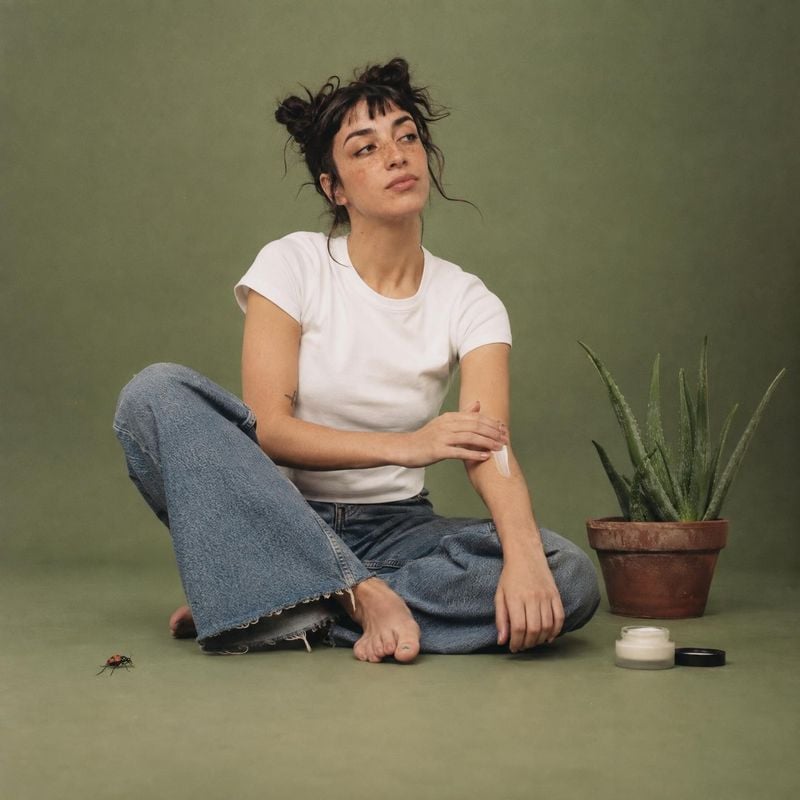 moisturizer cream model try-on product photo: A woman in a white T-shirt and jeans applies cream to her arm beside skincare jars and a potted aloe, posed baref…
