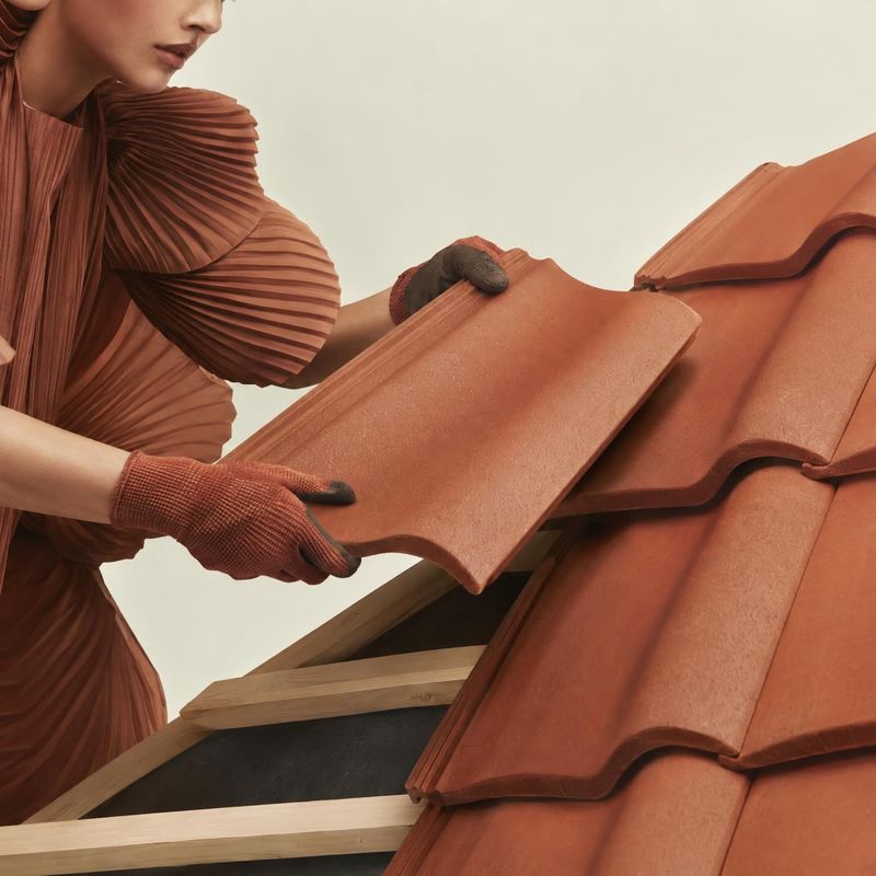roof tiles lifestyle scene product photo: A model in a sculptural rust-colored pleated outfit and gloves handles terracotta roof tiles against a pale studio ba…