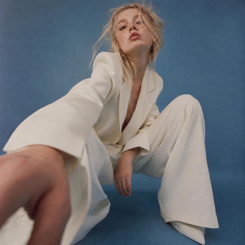 white tailored suit action & motion product photo: A blonde model poses low to the ground in a loose white tailored suit against a muted blue studio backdrop,…