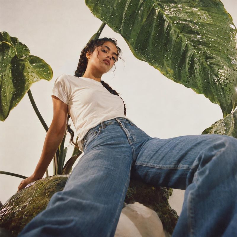 jeans model try-on product photo: A model in a white T-shirt and blue jeans poses from a low angle among broad rain-speckled leaves, with soft natural light an…