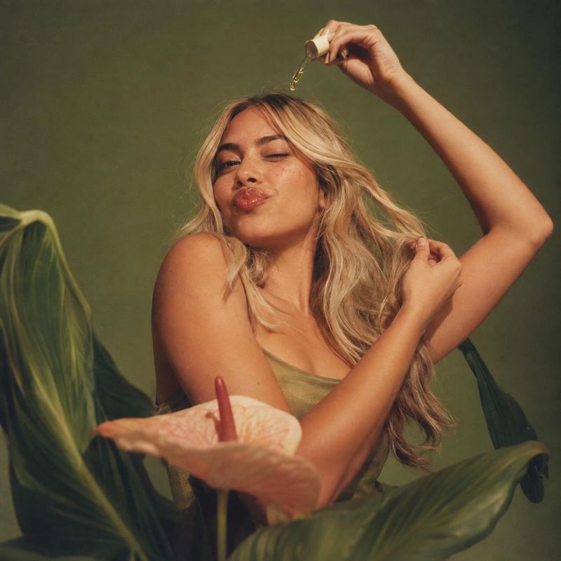 hair oil model try-on product photo: A blonde woman poses among lush tropical leaves, applying oil from a dropper above her hair in warm, soft studio light for…