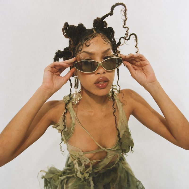 sunglasses model try-on product photo: A woman in sculptural braids and a distressed green outfit adjusts sleek oval sunglasses against a pale studio backdrop,…