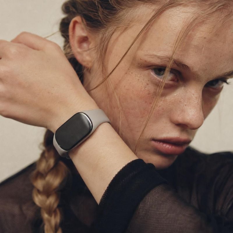 smartwatch model try-on product photo: A close, softly lit portrait shows a woman in a dark sheer-sleeved outfit wearing a sleek smartwatch, with a braided hai…