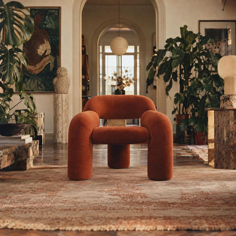 upholstered lounge chair room set product photo: A sculptural rust-orange upholstered chair sits on a patterned rug in a warm, art-filled interior with leafy p…