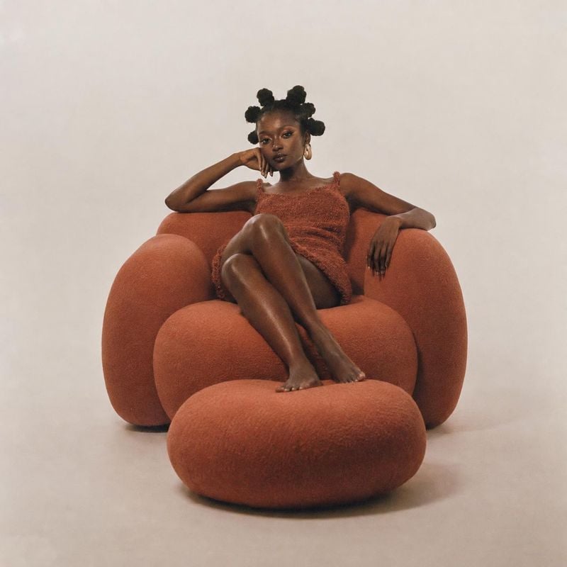 sculptural lounge chair and ottoman room set product photo: A model in a rust-toned textured dress sits barefoot on a sculptural rust-colored lounge chair and…