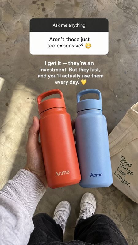 reusable water bottles product in hand product photo: A hand holds red and blue reusable bottles over a concrete floor, with casual shoes and a canvas tote nea…