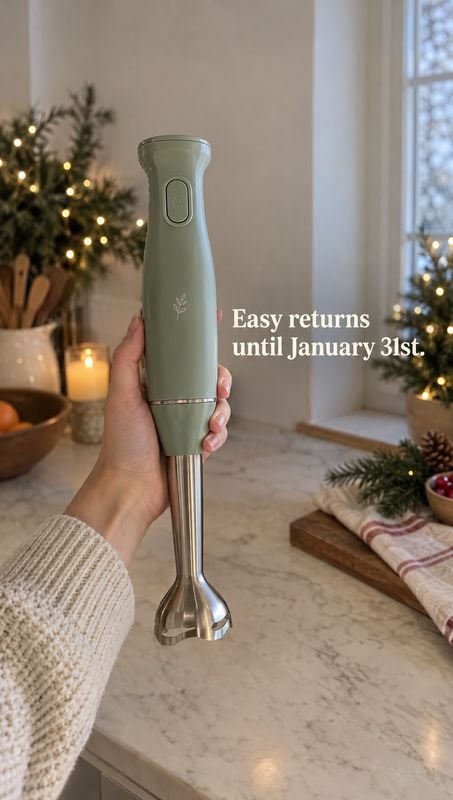 hand blender product in hand product photo: A hand presents a sage-green immersion blender over a marble kitchen counter, with warm candlelight, a softly lit h…