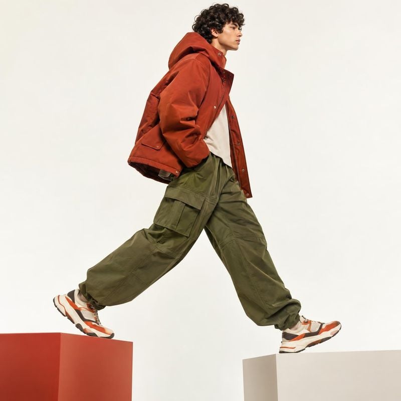 casual outfit and sneakers action & motion product photo: A curly-haired male model strides across two geometric blocks in a rust-red hooded jacket, olive carg…