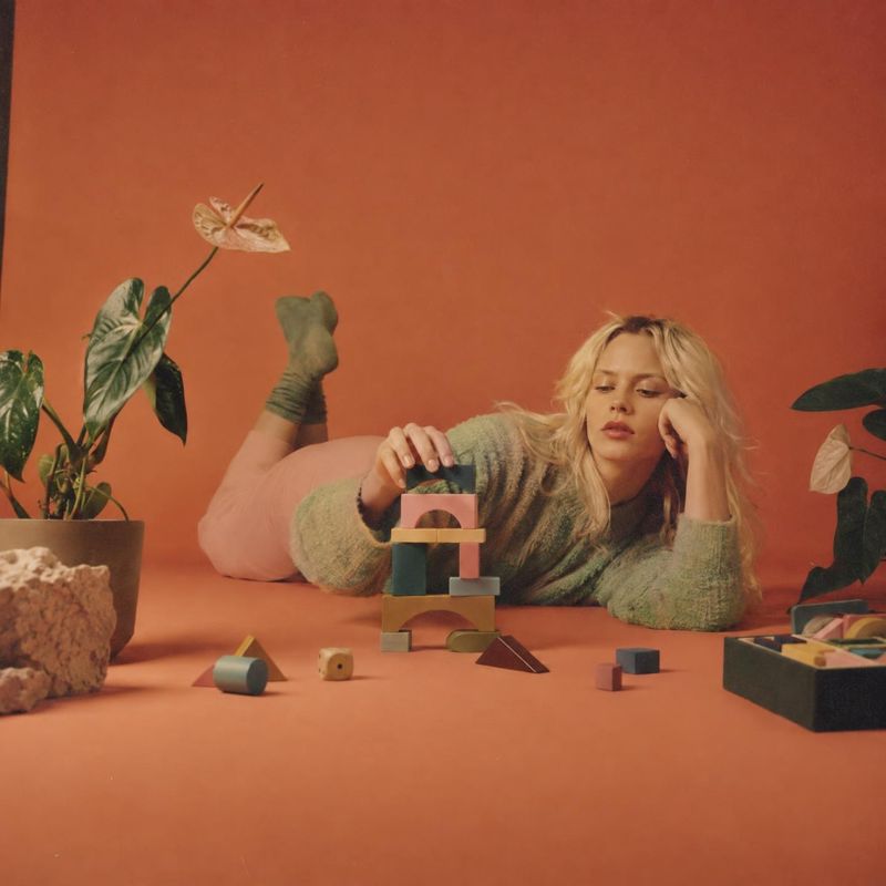 wooden building blocks lifestyle scene product photo: A blonde woman in a soft green sweater lounges among wooden building blocks and potted plants on a terrac…