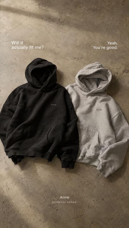 hoodies flat lay product photo: Two black and light-gray hoodies lie side by side on a textured concrete floor, with understated white copy and soft directiona…