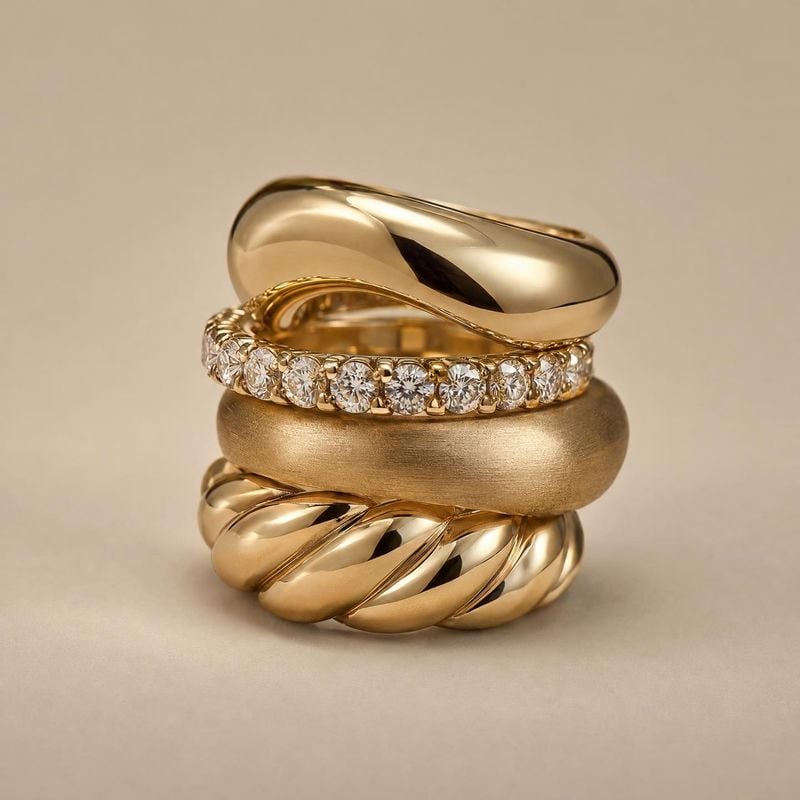 gold rings packshot product photo: A sculptural stack of polished and brushed gold rings, including a diamond-set band, is arranged on a warm beige studio surf…