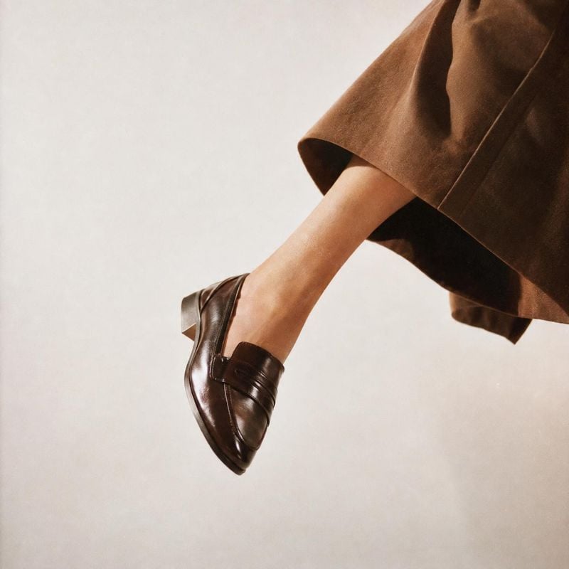 leather loafer model try-on product photo: A cropped model wears a polished dark leather loafer beneath a flowing brown skirt, suspended against a softly lit n…