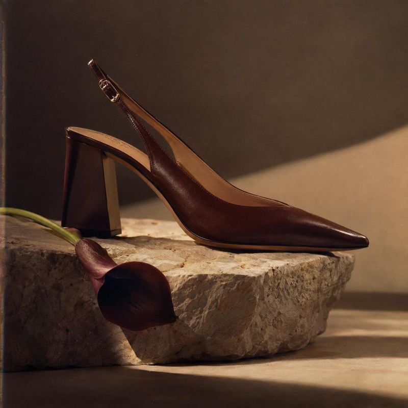 brown slingback heel fashion product photo: A polished dark brown slingback heel rests on a rugged stone plinth beside a sculptural flower, lit with warm direc…