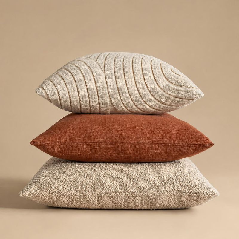 decorative pillows group lineup product photo: Three textured decorative pillows in cream and rust are stacked against a warm beige studio backdrop, with soft…