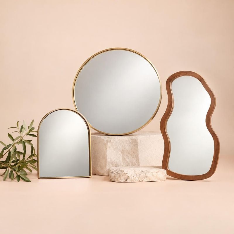 decorative mirrors group lineup product photo: Three mirrors in round, arched, and organic shapes are arranged on pale stone blocks against a warm blush backdr…