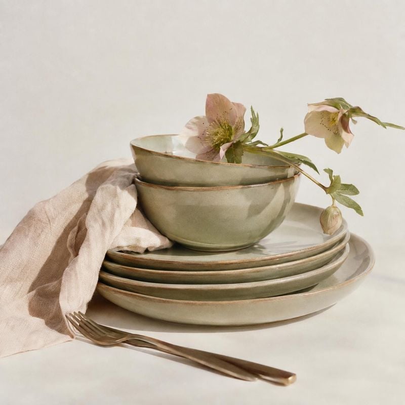 ceramic dinnerware set home product photo: A softly lit stack of muted green ceramic bowls and plates is styled with a linen napkin, fork, and delicate flowers…