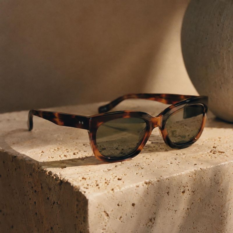 tortoiseshell sunglasses jewelry product photo: Tortoiseshell sunglasses rest on a textured stone block against a warm neutral backdrop, lit with soft directio…