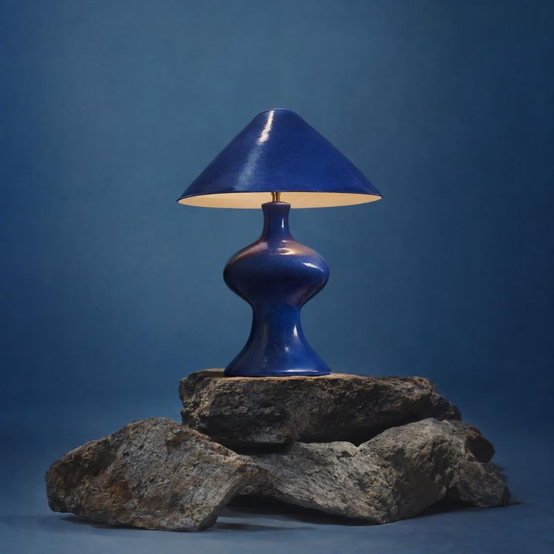 table lamp home product photo: A glossy cobalt-blue table lamp glows atop a rugged stack of rocks against a softly lit blue studio backdrop, creating a sculptu…