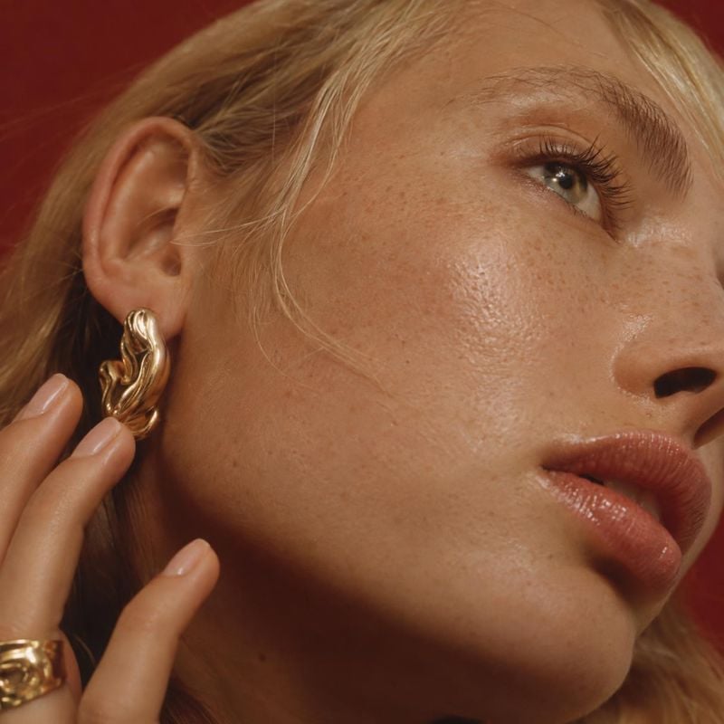 gold statement earrings beauty close-up product photo: A close-up portrait frames a woman wearing sculptural gold earrings and a matching ring against a deep r…