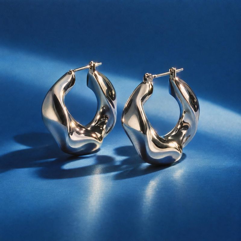 sculptural hoop earrings packshot product photo: A pair of polished sculptural silver-tone hoop earrings stands against a deep blue studio backdrop, with direc…