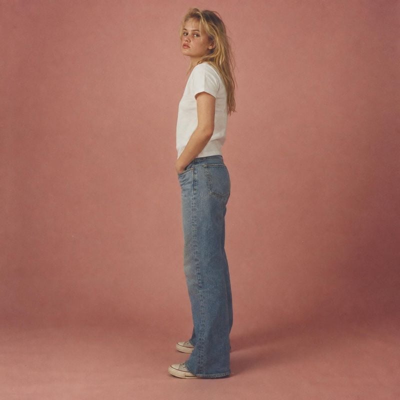 blue jeans model try-on product photo: A model poses in a white T-shirt, relaxed blue jeans, and sneakers against a muted pink studio backdrop, with soft light…