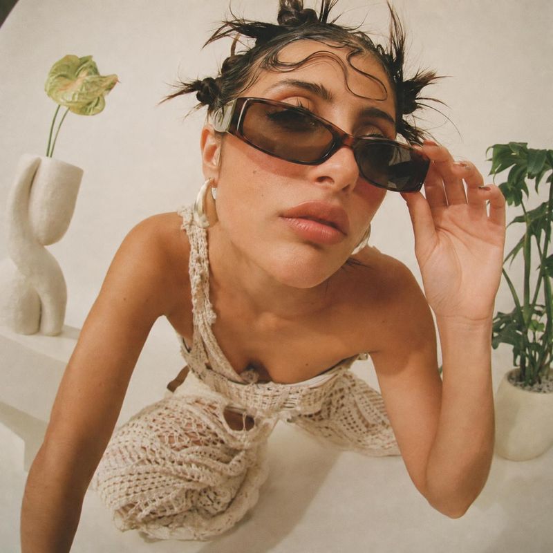 sunglasses model try-on product photo: A close, fashion-forward portrait shows a woman in dark sunglasses and a cream crochet outfit, adjusting the frames in a…