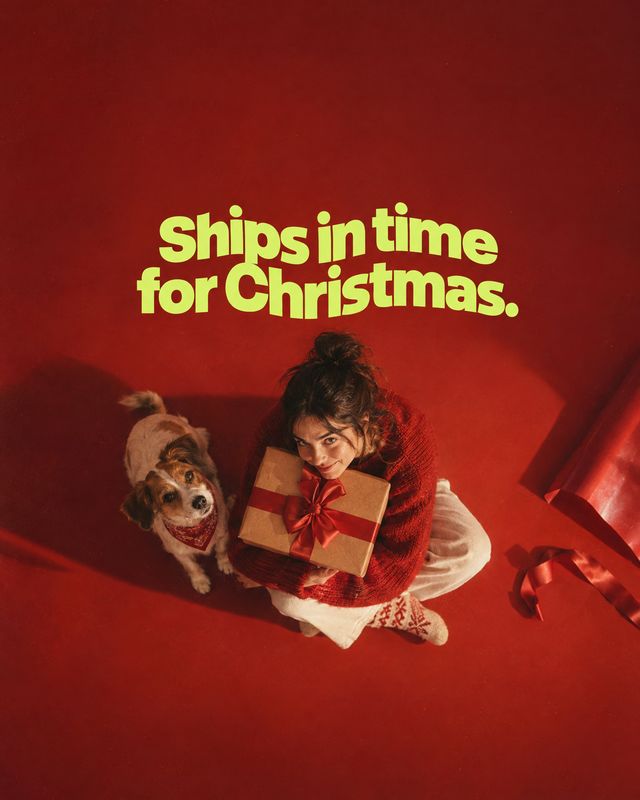 wrapped gift box christmas product photo: An overhead Christmas campaign scene shows a woman in a red sweater cuddling a ribbon-wrapped gift beside a small dog…