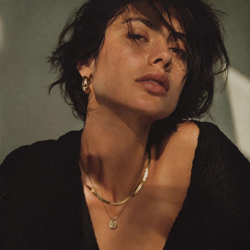 gold jewelry model try-on product photo: A close, warmly lit portrait shows a woman in a black top wearing layered gold necklaces and hoop earrings against a m…
