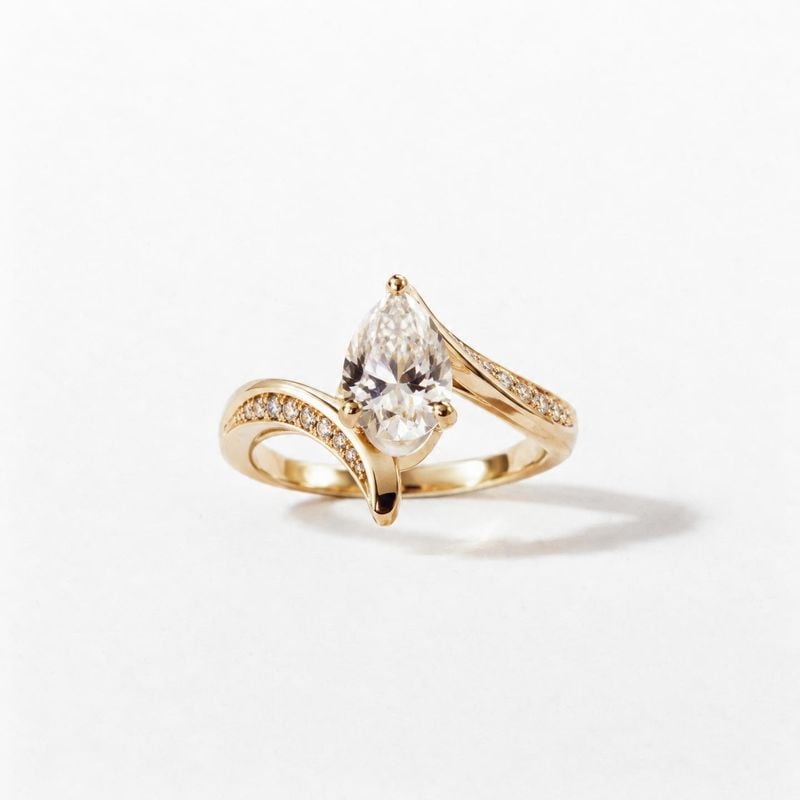 gold diamond ring packshot product photo: A sculptural gold ring with a prominent pear-shaped clear stone and pavé accents sits on a clean white surface, softl…