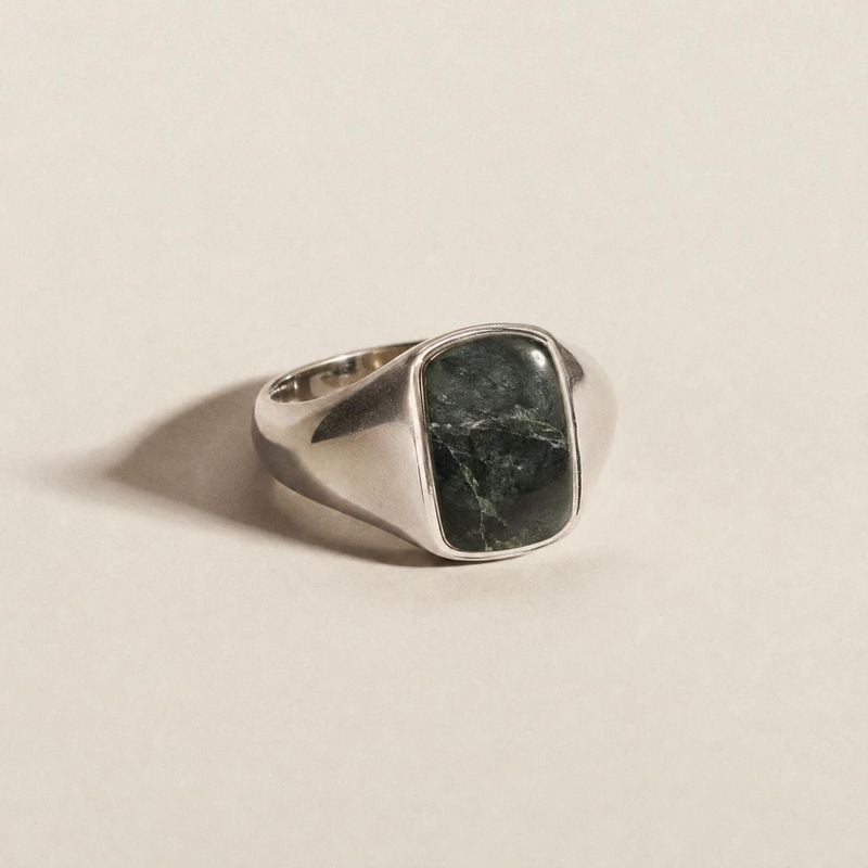 silver ring with dark stone packshot product photo: A silver signet-style ring with a dark green stone rests on a warm off-white surface, lit softly from the s…