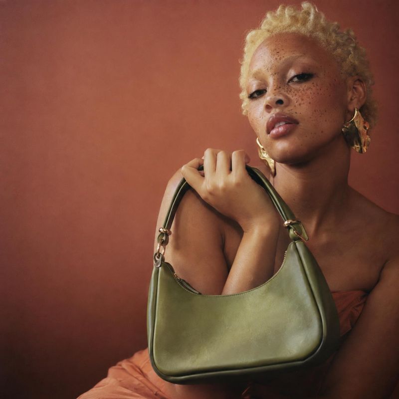 handbag model try-on product photo: A model poses with an olive-green shoulder bag against a warm terracotta studio backdrop, with soft directional light and a…