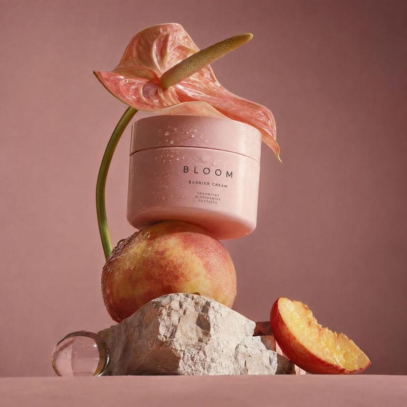 barrier cream jar ingredients product photo: A pink skincare jar is artfully balanced among a peach, a cut peach, pale stone, and a pink anthurium against a wa…