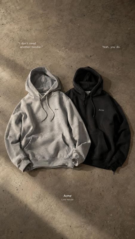 hoodies flat lay product photo: Two gray and black hoodies are arranged side by side on a textured concrete floor, with understated white copy and soft directi…