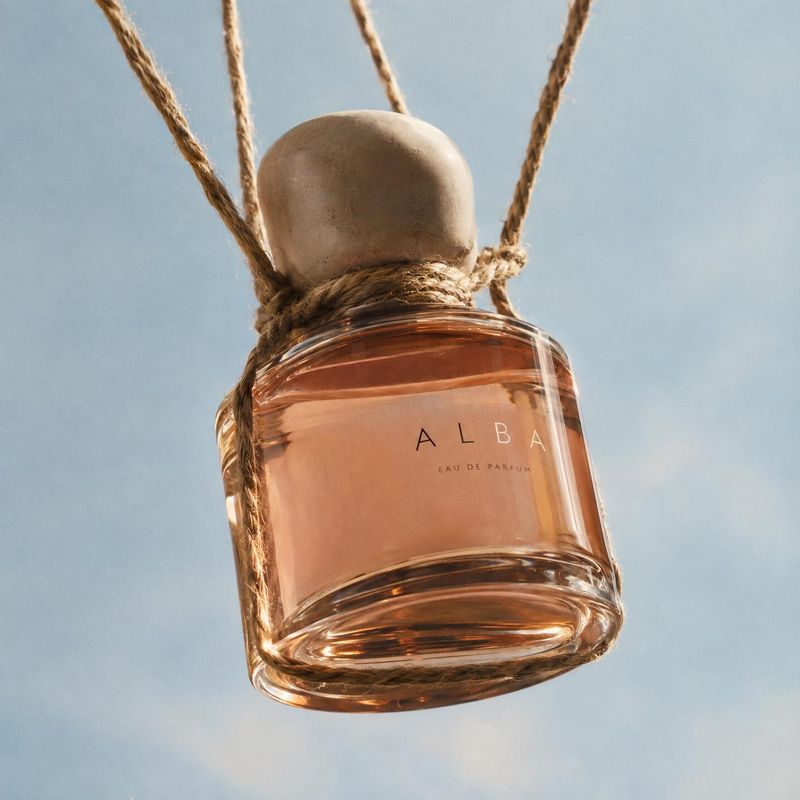 perfume floating product product photo: An amber perfume bottle labeled “ALBA” hangs from coarse ropes against a softly lit blue sky, creating an airy, warm-to…