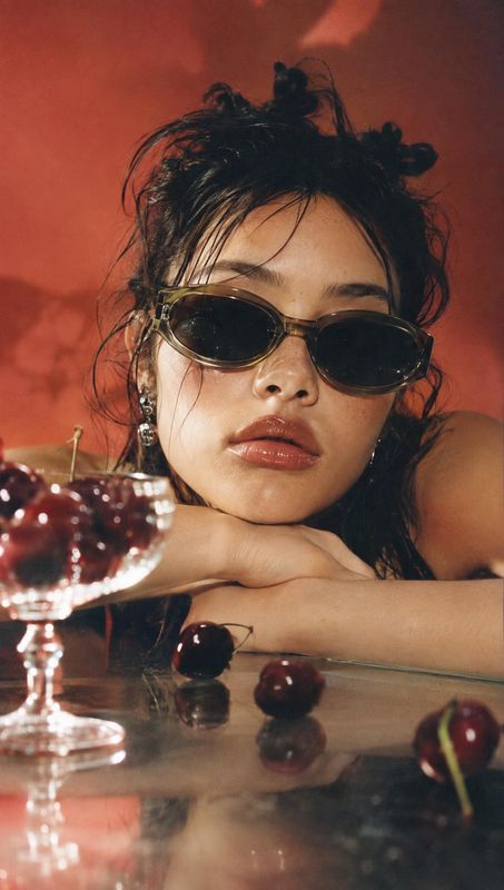 sunglasses model try-on product photo: A woman in sculptural sunglasses leans over a reflective table scattered with cherries, lit warmly against a rich red ba…