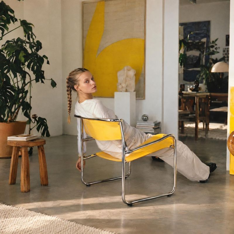 yellow lounge chair lifestyle scene product photo: A woman in a relaxed white outfit reclines in a bright yellow and chrome lounge chair in an art-filled inter…