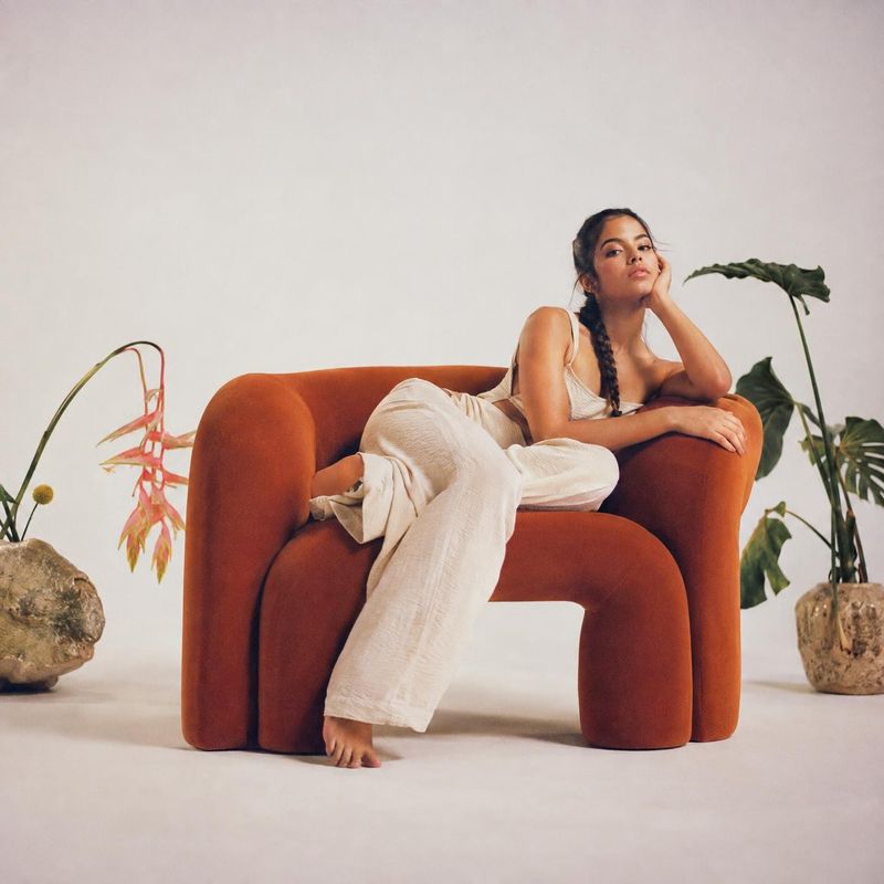 upholstered armchair room set product photo: A woman in a relaxed cream outfit reclines on a sculptural rust-colored armchair, styled with potted plants agains…