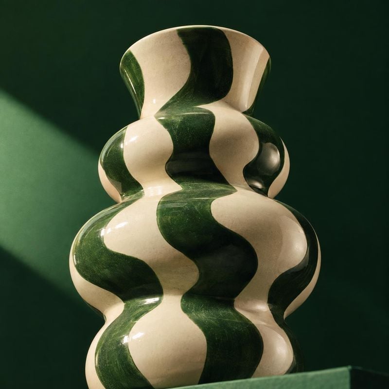 ceramic vase close-up detail product photo: A sculptural cream-and-deep-green striped ceramic vase fills the frame against a dark green studio backdrop, shaped…