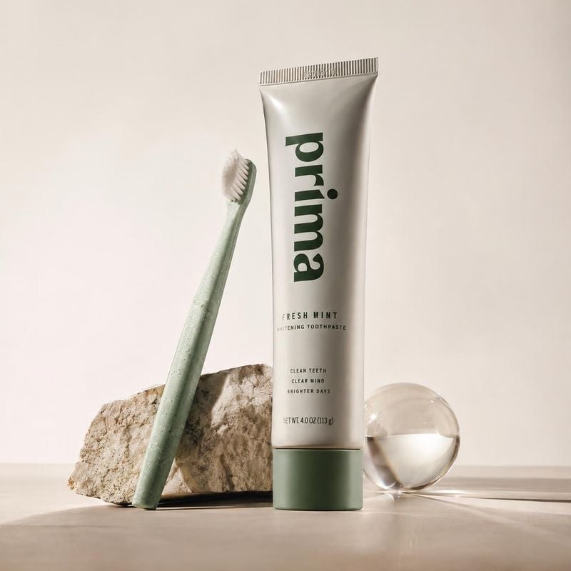 toothpaste personal care product photo: A toothpaste tube and pale green toothbrush stand among a stone fragment and glass sphere on a softly lit neutral studi…