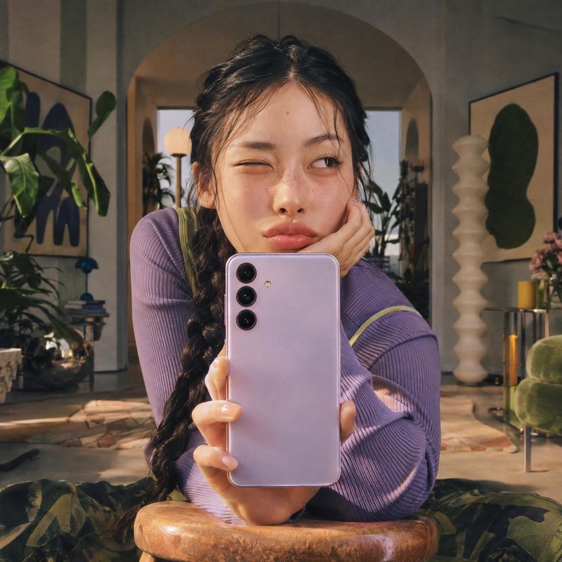 smartphone lifestyle scene product photo: A woman in a purple ribbed top holds a lavender smartphone toward the camera in a warmly lit, art-filled living room,…