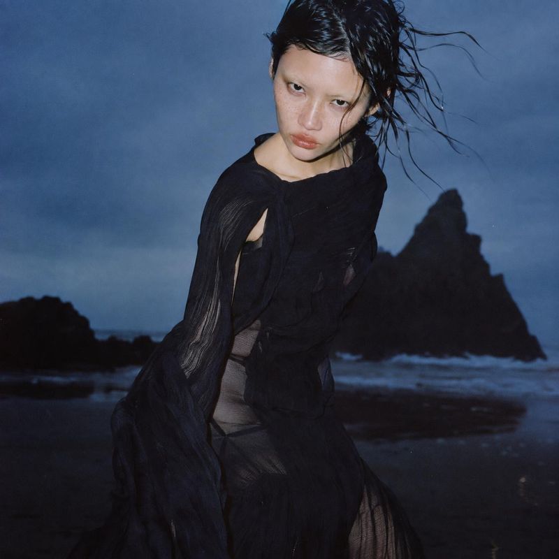 black sheer fashion outfit action & motion product photo: A model in a flowing black sheer outfit poses on a rocky shoreline, her windblown hair and cool blue…