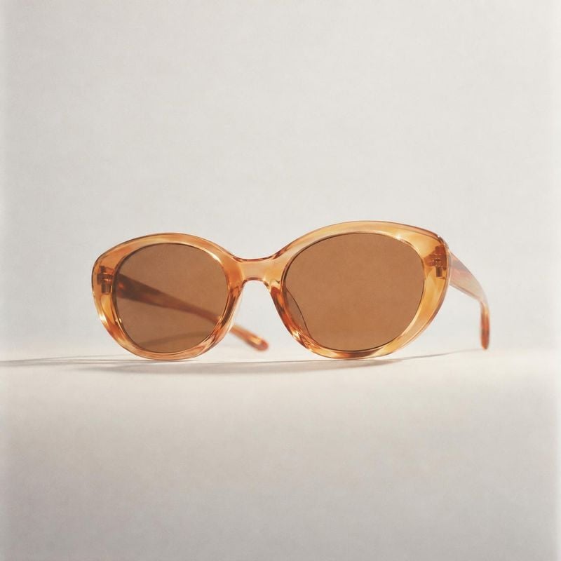 sunglasses jewelry product photo: Amber translucent oval sunglasses rest on a pale seamless surface, lit softly to bring out the warm frame and subtle shadow i…